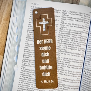 Könnte beinhalten: Brauner Leder-Lesezeichen mit dem weißen Text "DER HERR segne dich und behüte dich" und einem Kreuz-Design. Das Lesezeichen liegt auf einem offenen Buch.