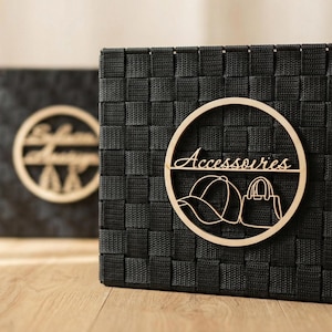 May include: Black woven storage basket with a wooden circular emblem that reads "Accessoires" and features a hat and handbag illustration. A second basket is visible in the background.