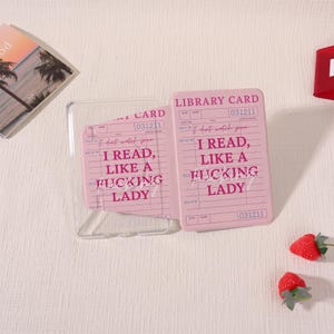 May include: Two pink phone cases with a library card design. The text on the cases reads "I READ, LIKE A FUCKING LADY". The cases are next to a clear phone case and two strawberries.