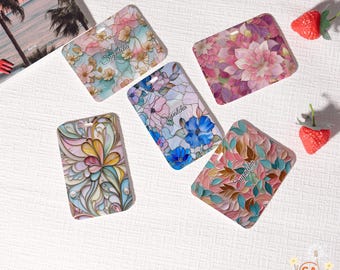 May include: Five rectangular luggage tags with floral designs in various colors. Each tag features a different pattern, including flowers and leaves, with the name "Supatcha" printed on them. The tags are arranged on a white surface.
