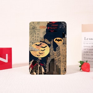 Könnte beinhalten: Ein rechteckiges Batman-Motiv mit einer Collage aus Comic-Panels. Das Design zeigt eine Stadtlandschaft, das Batman-Logo, Fledermäuse und die Wörter "POW" und "WHAAM!". Die Farbpalette ist eine Mischung aus Dunkelblau, Beige und Gelb.