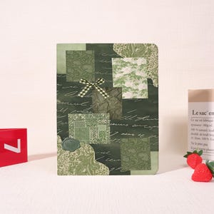 May include: A rectangular notebook with a dark green cover featuring a collage of various green patterned squares, cursive script text, a small green wax seal, and a decorative green and white checkered ribbon bow attached to the front center.