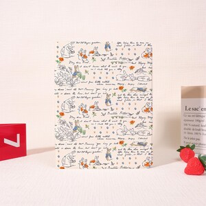 May include: A rectangular notepad featuring a repeating pattern of illustrations and text. The design includes drawings of rabbits, flowers, and handwritten script on a cream-colored background. The notepad is a charming stationery item.