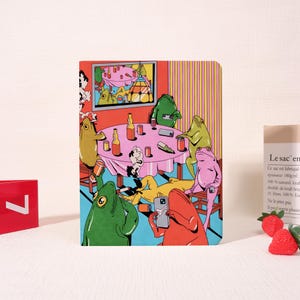 Könnte beinhalten: Ein farbenfrohes Notizbuch mit einer Cartoon-Illustration von Fröschen auf einer Party. Die Szene zeigt einen runden Tisch mit Flaschen und Telefonen, einen Fernseher und verschiedene Froschfiguren in unterschiedlichen Posen. Der Hintergrund hat vertikale Streifen.