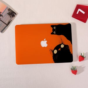 Peut inclure: Une coque rigide orange pour ordinateur portable ornée de deux illustrations de chats noirs aux yeux écarquillés. Un chat est placé en haut à droite et l'autre en bas à droite, encadrant le logo Apple blanc au centre de la coque.