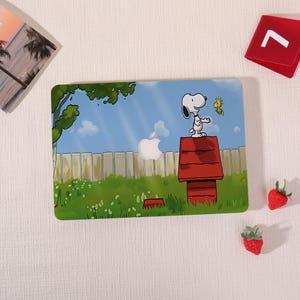 Könnte beinhalten: Ein Laptop-Skin mit einer Cartoon-Illustration von Snoopy, der auf einer roten Hundehütte steht, während Woodstock in der Nähe fliegt. Die Szene zeigt eine grüne Wiese, einen Holzzaun, einen blauen Himmel mit Wolken und eine Aussparung für das Apple-Logo.