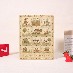 May include: A rectangular notebook with a cream-colored cover featuring a grid of twelve illustrations depicting farm life, including a person gardening, a house, sheep, cows, chickens, and a wheelbarrow, all framed by a floral and heart border.