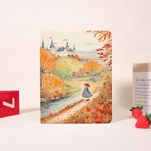May include: A notebook with a watercolour illustration of a girl in a blue dress walking towards a castle. The scene is set in autumn with orange and yellow trees and a river. The notebook has a white background.