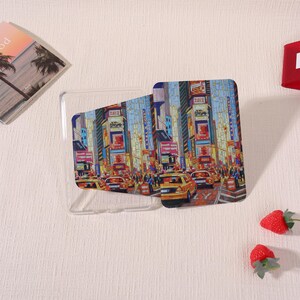 May include: A clear phone case with a colorful graphic of a city street scene, featuring yellow taxis, tall buildings, and bright signs. The case is shown with a matching insert. The image also includes a small book and strawberries.
