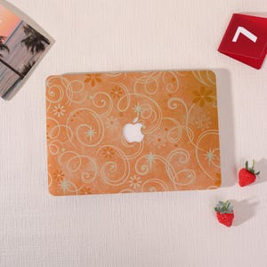 May include: A laptop hard shell case featuring an orange background with a repeating pattern of white swirling lines, small flowers, and star shapes. The center of the case has a cutout revealing the silver Apple logo on the laptop underneath.