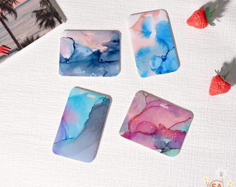 May include: Four phone cases with abstract watercolor designs in shades of blue, pink, and purple. One case is square, the others are rectangular with a hole at the top. Two red strawberry decorations are also present.