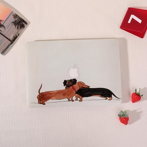May include: A clear laptop case featuring a watercolor illustration of two dachshund dogs. One dog is brown, the other is black, and both are wearing collars. The Apple logo is visible on the laptop.
