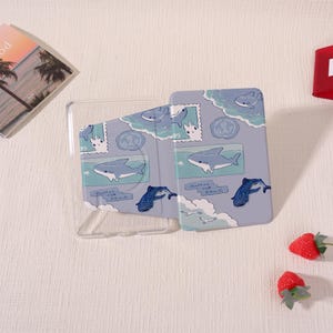 May include: A light blue phone case with a shark and whale design. The case has a clear plastic frame and a patterned cover featuring cartoon sharks, whales, and ocean elements. The design includes stamps and clouds.