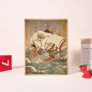 May include: A wooden print featuring a stylized illustration of a ship sailing on a stormy sea, with a skull and crossbones on the sail. The artwork is in a Japanese Ukiyo-e style, with text in Japanese. The print is rectangular and stands upright.