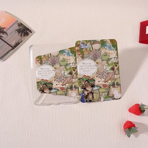 May include: Clear phone case with a whimsical design featuring fairies, gnomes, and floral elements. The case has the quote "What one loves in childhood stays in the heart forever." Two strawberries are on the right.