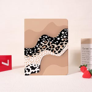 May include: A tan tablet case with a wavy design featuring black and white stars, leopard print, and cow print patterns. The case has a smooth finish and a neutral color palette.