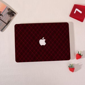 Könnte beinhalten: Eine weinrote Laptop-Hülle mit einem Karomuster und einem weißen Apple-Logo. Die rechteckige Hülle bedeckt die Oberseite eines Laptops. Zwei kleine Erdbeerdekorationen und ein rotes Quadrat mit einem weißen Pfeil sind ebenfalls vorhanden.