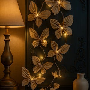 Op de afbeelding: Een decoratieve vlinderlichtslinger met warmwitte LED-lampjes. De vlinders zijn gemaakt van een getextureerd, beige materiaal en zijn geplaatst tegen een donkere achtergrond. Een vintage lamp en een kleine vaas zijn ook zichtbaar.