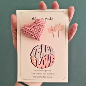 Peut inclure: Une carte avec un cœur rose crocheté et les mots "Peace & Love" dans une police rétro. La carte présente également de petites illustrations de cœurs et de signes de paix, avec du texte en français.