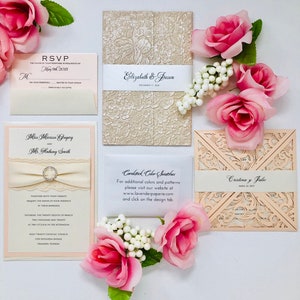 May include: Wedding invitation suite featuring various designs. Includes a cream-colored lace-patterned invitation, a peach-colored laser-cut invitation, and RSVP cards. Embellished with cream-colored ribbon, a rhinestone brooch, and pink roses.