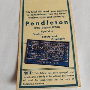May include: A rectangular cream-colored paper tag with dark teal borders and text, featuring a smaller, rectangular blue fabric label with gold embroidered text reading "Hand-Tailored from Pendleton Woolens, Portland, Oregon, 100% Virgin Wool, Made in U.S.A." centered on the card.