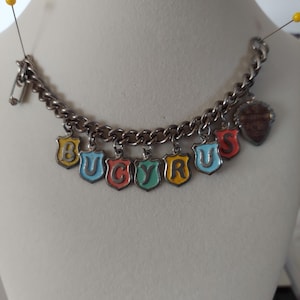 May include: A silver-toned metal charm bracelet featuring eight individual shield-shaped charms. Each charm displays a single letter in a different color, spelling out BUCYRUS, with an additional heart-shaped charm at the end.