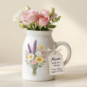Könnte beinhalten: Ein weißer Keramikkrug mit Henkel, gefüllt mit rosa und weißen Rosen. Der Krug ist mit einem Blumenstrauß-Design und einem Anhänger mit der Aufschrift "Muum With love always From Chloe, Sophie & Mia." verziert.