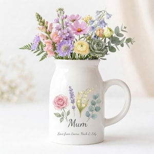 Könnte beinhalten: Eine weiße Keramikvase mit Henkel, gefüllt mit einem bunten Strauß frischer Blumen. Die Vase ist mit floralen Illustrationen und dem Wort "Mum" in Schreibschrift verziert, darunter eine persönliche Nachricht.