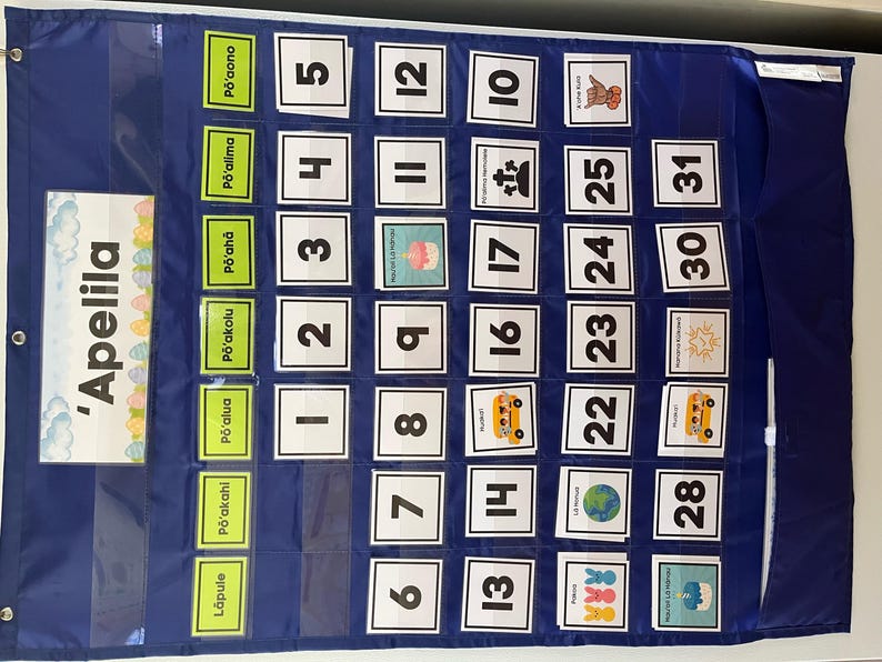 May include: A blue fabric calendar with pockets for each day of the month. The calendar has a title that says 'Apelila' and includes numbers 1 through 31. Each pocket has a different image, such as a school bus, a globe, and a birthday cake.