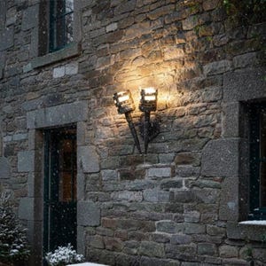 May include: Exterior wall light with two torch-shaped lamps. The lamps are black and emit a warm light. The fixture is mounted on a weathered stone wall, creating a rustic look. Snow is falling, enhancing the scene.