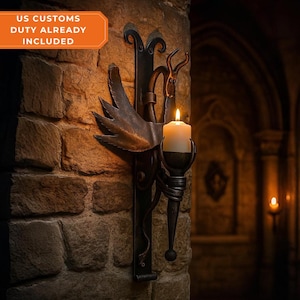 May include: A wrought iron wall sconce with a lit candle. The sconce features a wing-like design and decorative elements. The text "US CUSTOMS DUTY ALREADY INCLUDED" is in the upper left corner. The sconce is mounted on a stone wall.