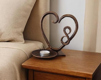May include: A dark bronze-colored metal sculpture shaped like a heart with an infinity symbol integrated into the design. The sculpture sits on a small, matching circular metal plate that holds a single white tea light candle.