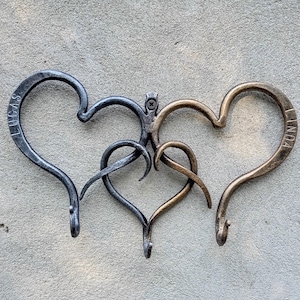 May include: Three intertwined metal heart-shaped hooks. One is dark gray with "LUCAS" inscribed, another is bronze, and the third is a darker bronze color with "LINDA" inscribed. The hooks are mounted on a gray surface.