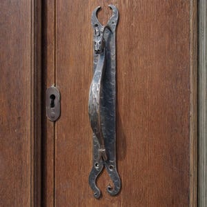 以下が含まれることがあります： 木製のドアに取り付けられた、ユニークなデザインの素朴な濃い金属製のドアハンドル。 ハンドルは、上部に様式化された動物の頭とテクスチャード加工されたグリップを備えています。 ドアには鍵穴もあります。 ドアハンドルは約30cmです。