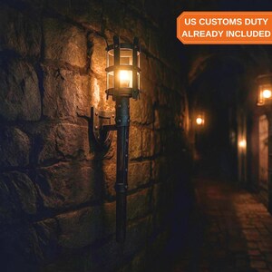May include: A wall-mounted torch-style light fixture featuring a dark metal frame with a cylindrical cage top and a warm glowing light bulb. The fixture is attached to a textured stone wall, casting a soft illumination across the surface.