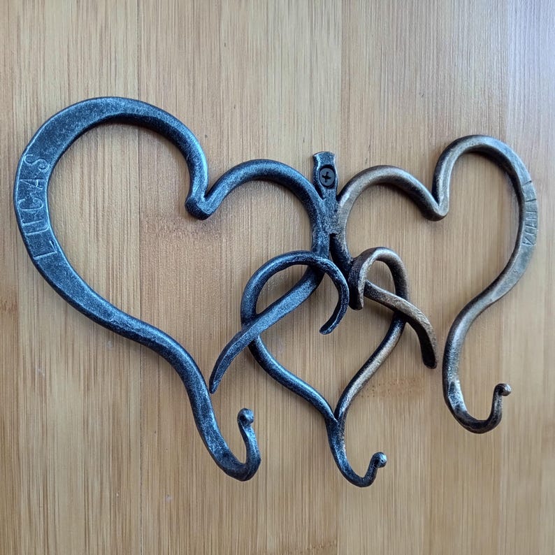 May include: Hand-forged metal wall hook with a heart design. The hook features three intertwined hearts in shades of gray and bronze, with the names "LUCAS" and "KAIA" inscribed on two of the hearts. The hooks are mounted on a wooden surface.