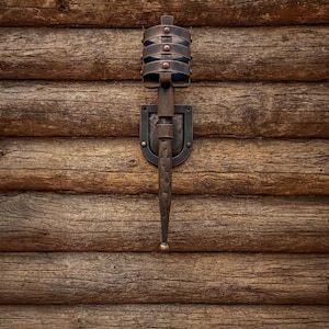 May include: A metal wall-mounted torch holder with a dark, aged finish. The torch has a cylindrical cage design with horizontal bands and a decorative shield-shaped base. It's mounted on a weathered wooden wall, creating a vintage aesthetic. The torch holder is made of metal.