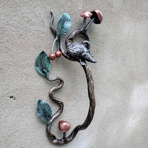 Könnte beinhalten: Dekorativer Türgriff aus Metall mit einem stilisierten Igel auf einem Ast mit Blättern, Kirschen und Pilzen. Der Griff ist bronzefarben mit grünen und kupferfarbenen Akzenten, montiert auf einer strukturierten grauen Oberfläche.