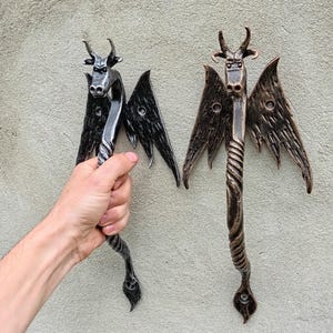 May include: Two handcrafted metal door handles, each featuring a stylized dragon design with horns, wings, and a twisted handle. One is held by a hand. The handles are finished in shades of black and bronze, adding a unique, artistic touch to any door.