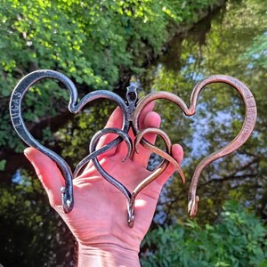May include: Hand-forged metal sculpture featuring three intertwined hearts in shades of silver, bronze, and copper. The silver heart is inscribed with the name "LUCAS", while the copper heart has "AMMA". The sculpture has a unique, artistic design.