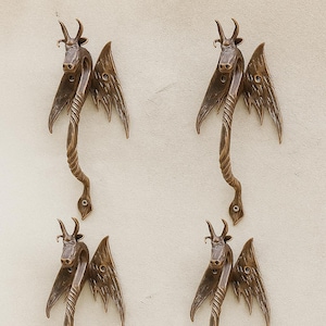 Könnte beinhalten: Vier bronzefarbene Türgriffe mit einem einzigartigen Drachendesign. Jeder Griff hat einen Drachenkopf mit Hörnern, Flügeln und einem geschwungenen Körper. An einer hellen Wand montiert.