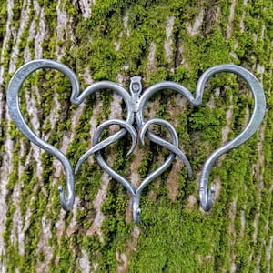 May include: A handcrafted metal wall hook featuring a heart design. The hook is made of silver-colored metal and has three hooks. The design is mounted on a tree trunk covered in green moss.