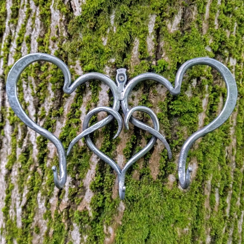 May include: A handcrafted metal wall hook featuring a heart design. The hook is made of silver-colored metal and has three hooks. The design is mounted on a tree trunk covered in green moss.