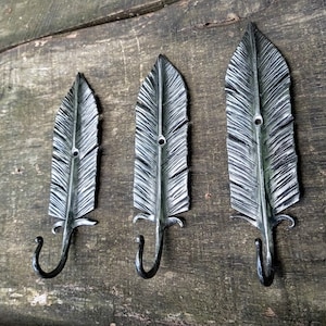 May include: Three metal feather-shaped wall hooks. The hooks are silver with black accents and a curved hook at the bottom. The feathers have detailed veining and are mounted on a wooden surface.