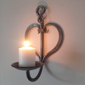 May include: A handcrafted, metal wall sconce in the shape of a heart, holding a lit white candle. The sconce has a dark, rustic finish and features the words "6 Happy Years" etched into the heart shape. The candleholder is a small, round dish.