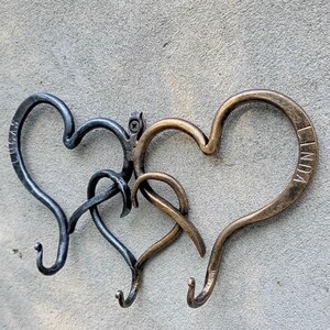 May include: Three metal heart-shaped wall hooks. One is dark gray with the name "LUCAS" on it, another is a lighter gray, and the third is bronze with the name "LINDA". The hooks are mounted on a gray textured wall.