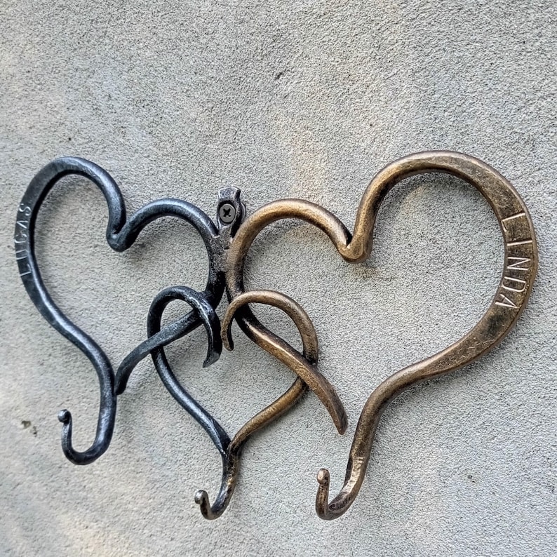 May include: Three metal heart-shaped wall hooks. One is dark gray with the name "LUCAS" on it, another is a lighter gray, and the third is bronze with the name "LINDA". The hooks are mounted on a gray textured wall.