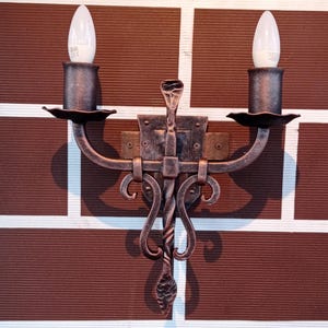 May include: A wrought iron wall sconce with two candle-shaped light bulbs. The sconce has a dark brown finish with decorative scrollwork and a central vertical element. The sconce is mounted on a brown brick wall.