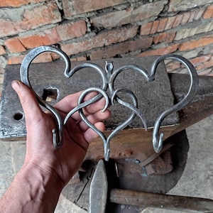 May include: Hand-forged metal wall art featuring three interconnected hearts. The piece is crafted from iron and has a rustic, industrial aesthetic. The hearts are connected by a butterfly-like design. The hooks are perfect for hanging keys or small items.