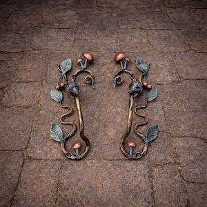 Könnte beinhalten: Zwei verzierte Türgriffe aus Metall in Bronzefarbe. Jeder Griff hat ein skurriles Design mit Blättern, Pilzen und einer kleinen Tierfigur. Die Griffe sind auf einer strukturierten, braunen Steinoberfläche angeordnet.